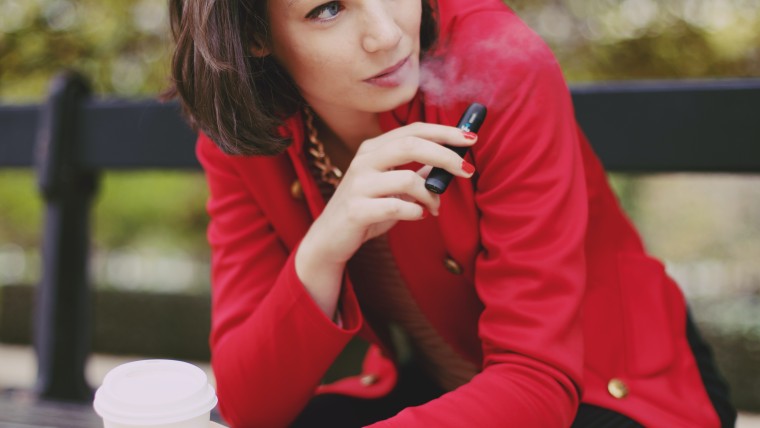 Woman sitting outside with a red blazer and black pants drinking a coffee and vaping.